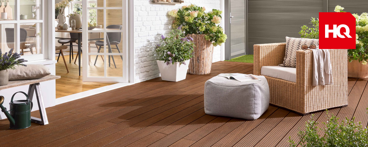 Holzfarbene Terrasse mit einem Flechtsessel und Pouf sowie bepflanzten Kübeln vor einer weißen Hauswand.